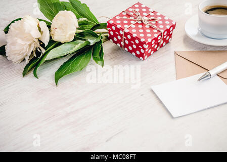 Red dotted gift box, kraft envelope and empty greeting card, coffee cup and peony flowers bouquet over white wood background. Copy space. Stock Photo