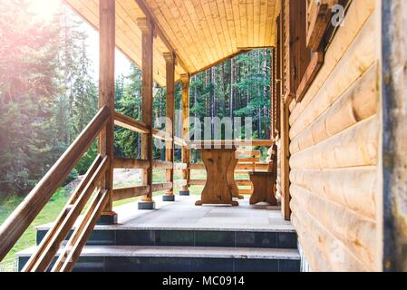 Side view of an open veranda in front of a wooden forest cottage. Pine forest under the sun rays in the background Stock Photo