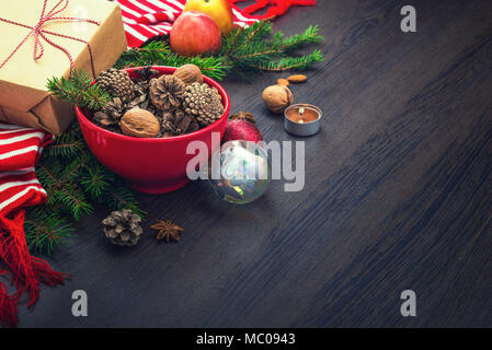 Christmas decoration - red bowl full of fir-cones, gift box wrapped in kraft paper, pine branches, candle, nuts, anise, apples, christmas toys and red Stock Photo