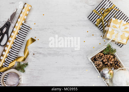 Gift box wrapped in black and white striped paper with golden ribbon, a crate full of pine cones and christmas toys and wrapping materials on a white  Stock Photo