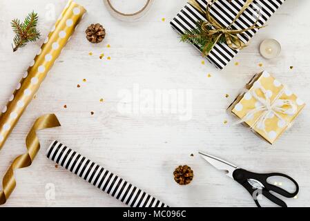 Gift boxes wrapped in black and white striped and goden dotted paper with, pine, cones, candle and wrapping materials on a white wood old background.  Stock Photo