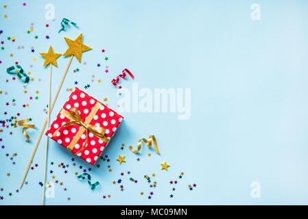 Top view of a red dotted gift box, golden magic wands, colorful confetti and ribbons over blue background. Celebration concept. Copy space. Stock Photo