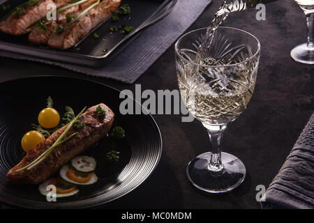 Portion of delicious roasted salmon on black plate with fresh vegetables and herbs on the stylish black table. White wine is pouring in clean crystal  Stock Photo