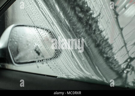 View from inside of car to side mirror and window being washed in carwash, brush leaving strokes in soap foam and shampoo. Stock Photo