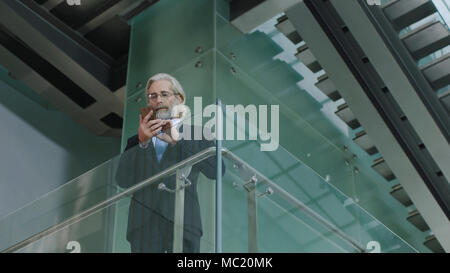senior corporate executive standing on second floor in modern office building dialing numbers using cellphone. Stock Photo