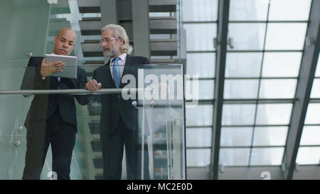 latino and caucasian corporate executives standing on second floor of modern building discussing business using digital tablet. Stock Photo