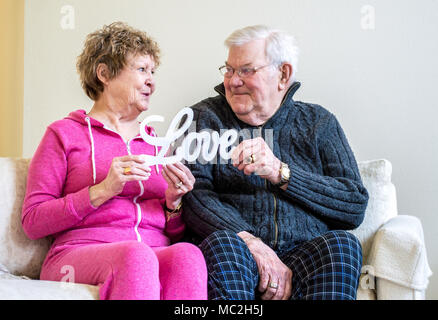 mature Caucasian couple smiling at each other and holding a wooden sign saying 'Love' while sitting on a sofa Stock Photo