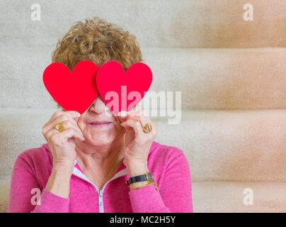 medium shot of a smiling middle aged Caucasian woman holding red paper hearts over her eyes while sitting on carpeted stairs Stock Photo