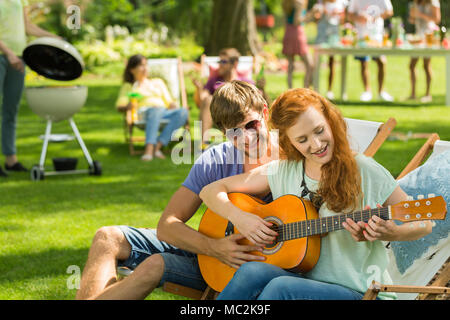 Young boy teaches playing guitar redheaded girlfriend while meeting with friends Stock Photo