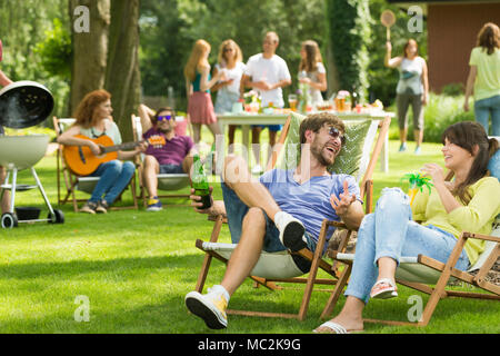 Friends sit on sunbeds and drink drinks while barbecuing in the garden with friends Stock Photo