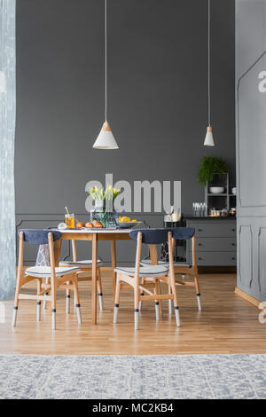 Low hanging pendant light above a small, wooden kitchen table with flowers in a monochromatic dining room interior with molding on tall gray walls Stock Photo