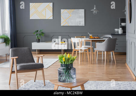 Yellow tulips and a modern gray armchair in a stylish, multifunctional living room interior with abstract paintings and designer dining chairs Stock Photo