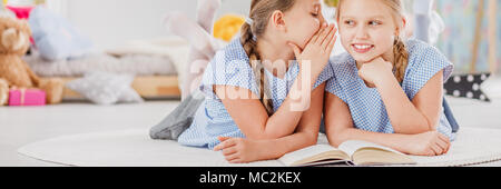 Young girl whispering secrets to her best friend while lying on the floor in children's bedroom and reading a book together Stock Photo