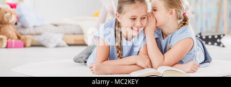 Happy young girl whispering a funny joke to her sister while spending time together in a bright kid's bedroom Stock Photo