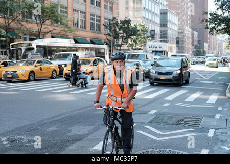 Young male cyclist seen wearing a high vis jacket and bike helmet, waiting at a busy junction with traffic behind him near a crossing area. Stock Photo