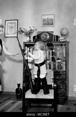 Mom helps her daughter become a little German soldier complete with gun, pipe, boots and dagger, ca. 1938. Stock Photo