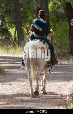 Warrior in Comanche clothing riding white horse along a trail through the forest Stock Photo