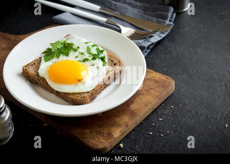 Fried Egg on Toast  for Breakfast. Fried egg with bread and parsley on white plate on black, top view, copy space. Stock Photo