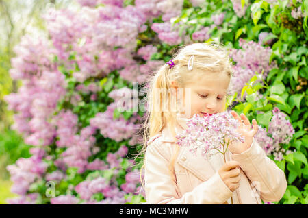 adorable blond girl holding lilac flowers and smelling to them outdoors next to blooming lilac bush Stock Photo