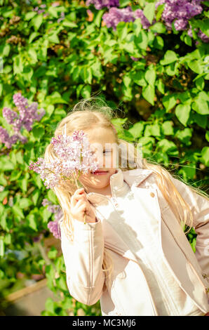 adorable blond girl holding lilac flowers and smelling to them outdoors next to blooming lilac bush Stock Photo