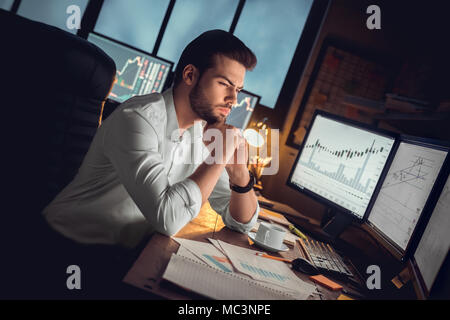 Focused thoughtful trader or serious investor working at night overtime analyzing stock trading graphs looking at computer monitors controlling market