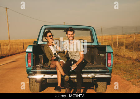Smiling couple sitting in the back of their pickup truck enjoying the road trip in country side. Man and woman holding a bottle of drink enjoying the Stock Photo