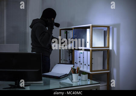 Thief Wearing Balaclava Stealing File From Shelf At Workplace Stock Photo