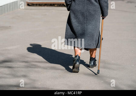 On a sunny day a old woman walking down the street with walking stick. The woman's shadow is visible on the sidewalk. View from the back. Stock Photo