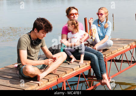 Family spending vacation time together having a snack sitting on jetty over the lake on sunny day in the summertime. Quality family time at weekend Stock Photo