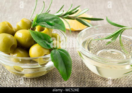 Green olives with green leaves and rosemary in a small glass cup on an old table cloth Stock Photo