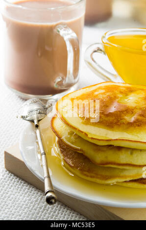 Glass mug with cocoa, silver spoon, honey and pancakes for dessert closeup with shallow depth of field. Stock Photo