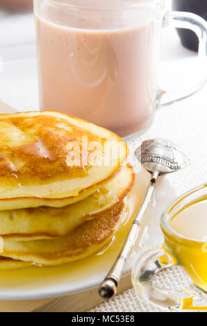 Glass mug with cocoa, silver spoon, honey and pancakes for dessert closeup with shallow depth of field. Stock Photo
