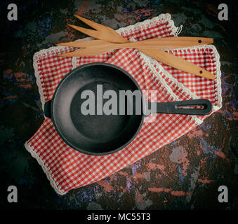 a round empty black cast-iron frying pan on a red napkin in a red box on a brown worn background, next to two wooden forks Stock Photo
