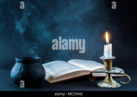 Candle in candlestick burning and smoky pot and old open book on dark and smoked background. Halloween concept Stock Photo