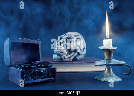 Mystical box with black rock and candle in candlestick burning and skull on book blurry in background, smoked and dark scene. Halloween concept. Stock Photo