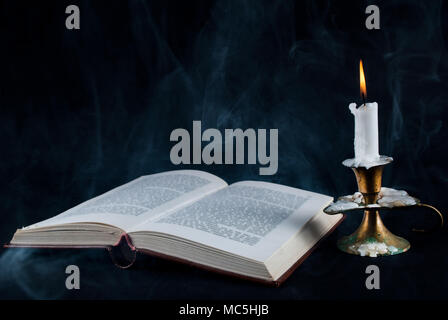 Old open book and candle in candlestick and smoke on dark black background Stock Photo