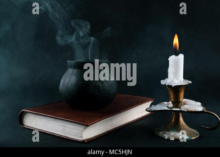 Wicker pot is smoked on the old book next to candle in old candlestick black and smoked background. Halloween concept Stock Photo