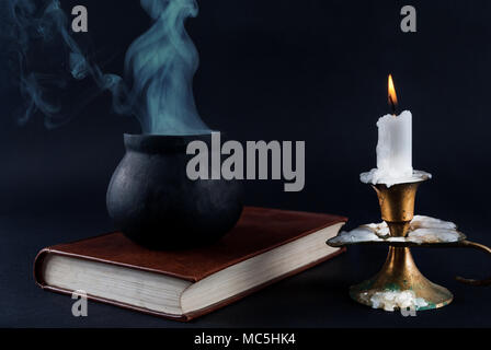 Wicker pot is smoked on the old book next to candle in old candlestick isolated on black. Halloween concept Stock Photo