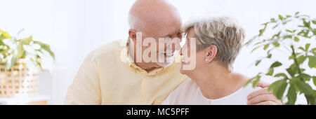 A close-up portrait of a happy senior couple - an older caucasian man and woman embracing face to face, touching noses and smiling in a white bright r Stock Photo