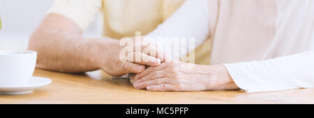 A close-up of wrinkled arms on a table, an older man holding his hand on an elderly woman's hands showing affection and supporting her Stock Photo