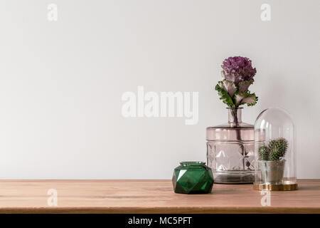 Close-up of a flower in a pink glass vase and a small cactus in a dome on the side of a wooden surface and an empty, white background Stock Photo