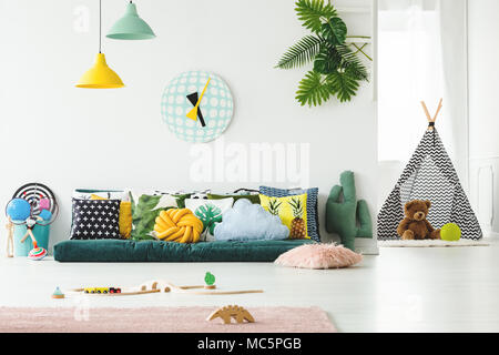 Dark green sofa for children with decorative cushions placed in white room interior with toys and patterned teepee Stock Photo