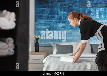 A young, working maid laying fresh, white towels on a bed in a hotel