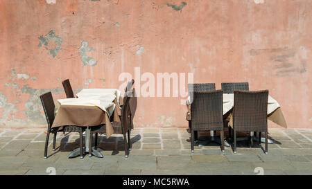 Two outdoor restaurant tables with tablecloth and chairs against a dilapidated and cracked wall on bright sunshine Stock Photo