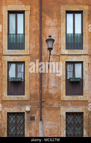 A weathered facade to an apartment block in Rome, Italy, in typical earth tones with a street lamp on the wall and windows some with boxes but no plan Stock Photo