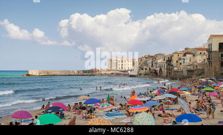 Cefalu, Sicily, Italy, crowds of people cover the sandy beach enjoying their holiday on a bright sunny August day with the old town buildings as a bac Stock Photo