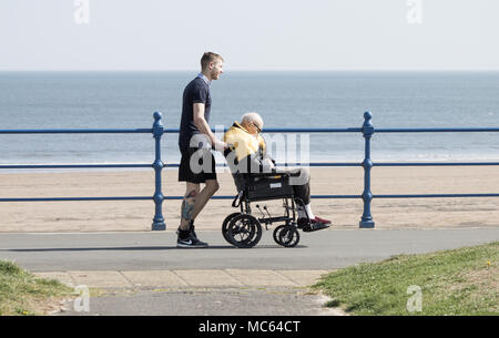Young man (possibly carer) pushing elderly man in wheelchair at coast. UK Stock Photo