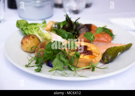 Delicious buffet lunch of tartlet, smoked salmon, new potatoes, salad greens, micro herbs and gherkin on a white plate and white tablecloth. Stock Photo