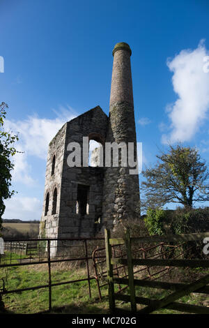 Cornish tin mine showing engine house and stack against a bright blue sky with clouds on a sunny day with old iron fence and trees in the foreground. Stock Photo