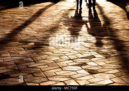 Blurry long shadows silhouettes of a couple walking on city patterned square in sunset Stock Photo